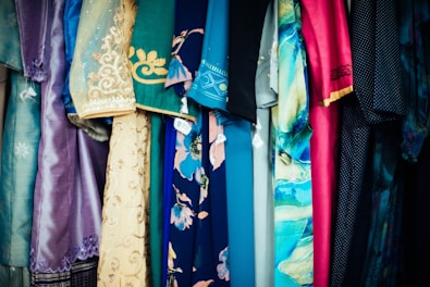 A variety of colorful fabrics and garments hang together, showcasing intricate patterns and a mix of textures. The textiles include floral designs, geometric patterns, and embroidery, featuring shades of blue, pink, gold, and teal.
