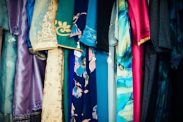 A variety of colorful fabrics and garments hang together, showcasing intricate patterns and a mix of textures. The textiles include floral designs, geometric patterns, and embroidery, featuring shades of blue, pink, gold, and teal.