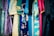 A variety of colorful fabrics and garments hang together, showcasing intricate patterns and a mix of textures. The textiles include floral designs, geometric patterns, and embroidery, featuring shades of blue, pink, gold, and teal.