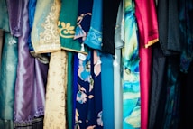 A variety of colorful fabrics and garments hang together, showcasing intricate patterns and a mix of textures. The textiles include floral designs, geometric patterns, and embroidery, featuring shades of blue, pink, gold, and teal.