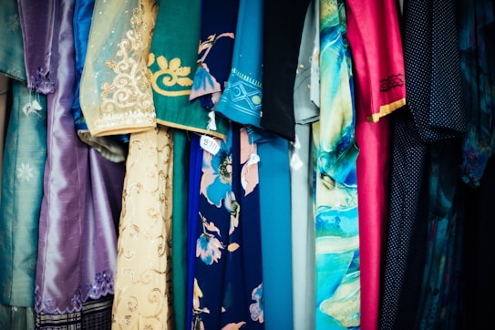 A variety of colorful fabrics and garments hang together, showcasing intricate patterns and a mix of textures. The textiles include floral designs, geometric patterns, and embroidery, featuring shades of blue, pink, gold, and teal.