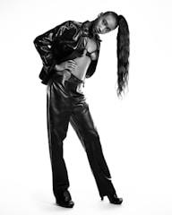 A person poses in a stylish leather outfit consisting of a jacket and pants. The outfit is accessorized with high heels and the hair is styled in a long, sleek ponytail. The individual strikes a confident pose, standing slightly tilted against a white background.