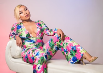 A person is reclining on a white, rounded-edge sofa wearing a vibrant floral jumpsuit. The fabric features bold colors like pink, green, and blue, with a deep V-neckline. Their blonde hair is styled to the side, and they are wearing light makeup. The background has a subtle gradient transitioning from pink to blue.