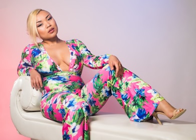 A person is reclining on a white, rounded-edge sofa wearing a vibrant floral jumpsuit. The fabric features bold colors like pink, green, and blue, with a deep V-neckline. Their blonde hair is styled to the side, and they are wearing light makeup. The background has a subtle gradient transitioning from pink to blue.