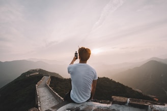 man sitting on cliff facing mountains
