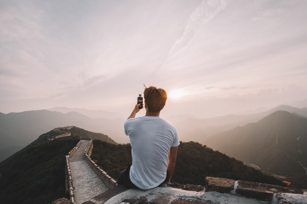 man sitting on cliff facing mountains