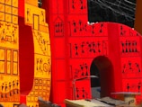 Large, vibrant letters adorned with intricate tribal-style illustrations. The design features small human figures engaged in various activities, constructed in a traditional, artistic pattern. The letters are primarily in shades of red and yellow, with black line drawings that depict scenes and motifs inspired by indigenous art.