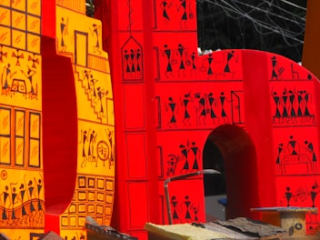 Large, vibrant letters adorned with intricate tribal-style illustrations. The design features small human figures engaged in various activities, constructed in a traditional, artistic pattern. The letters are primarily in shades of red and yellow, with black line drawings that depict scenes and motifs inspired by indigenous art.