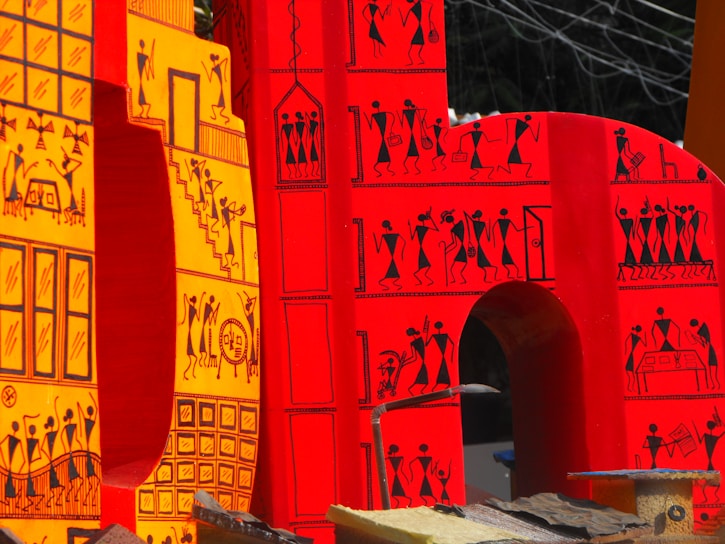 Large, vibrant letters adorned with intricate tribal-style illustrations. The design features small human figures engaged in various activities, constructed in a traditional, artistic pattern. The letters are primarily in shades of red and yellow, with black line drawings that depict scenes and motifs inspired by indigenous art.