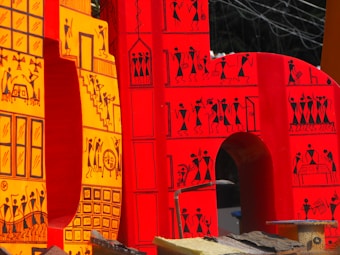 Large, vibrant letters adorned with intricate tribal-style illustrations. The design features small human figures engaged in various activities, constructed in a traditional, artistic pattern. The letters are primarily in shades of red and yellow, with black line drawings that depict scenes and motifs inspired by indigenous art.