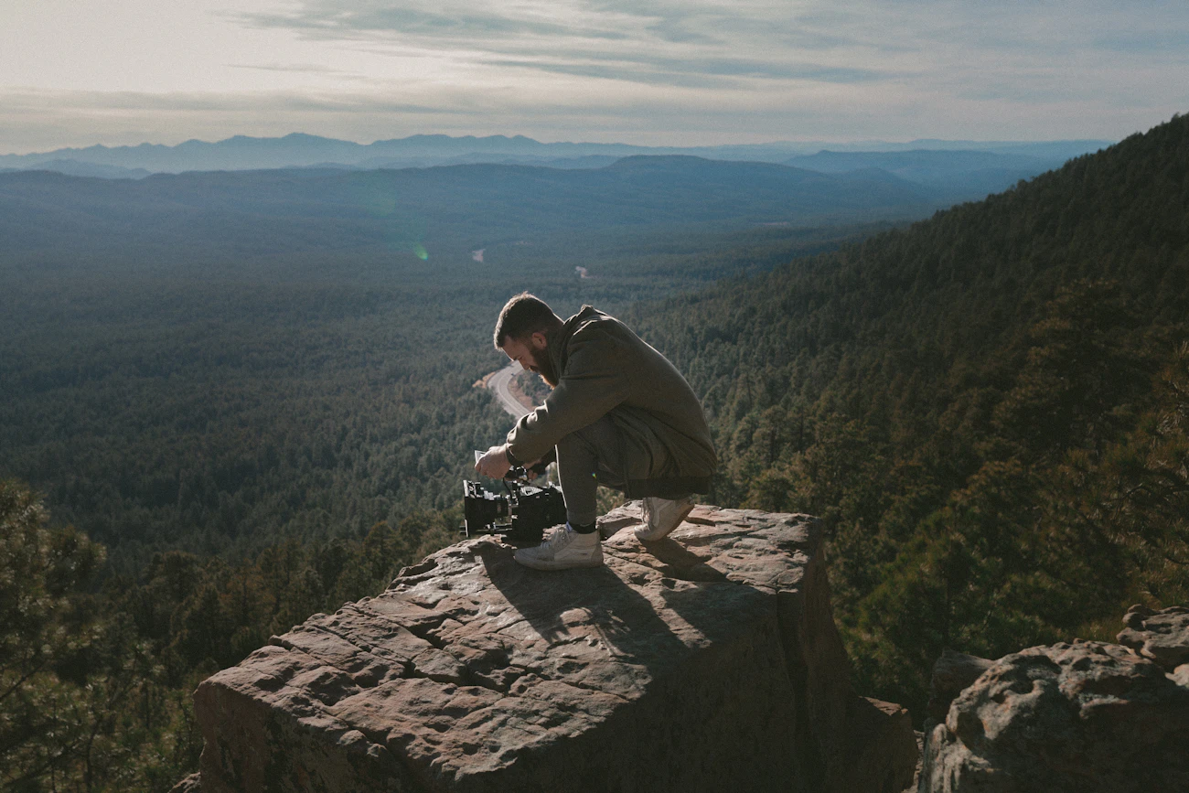 Camera operator crouching on a rocky cliff above a forested valley