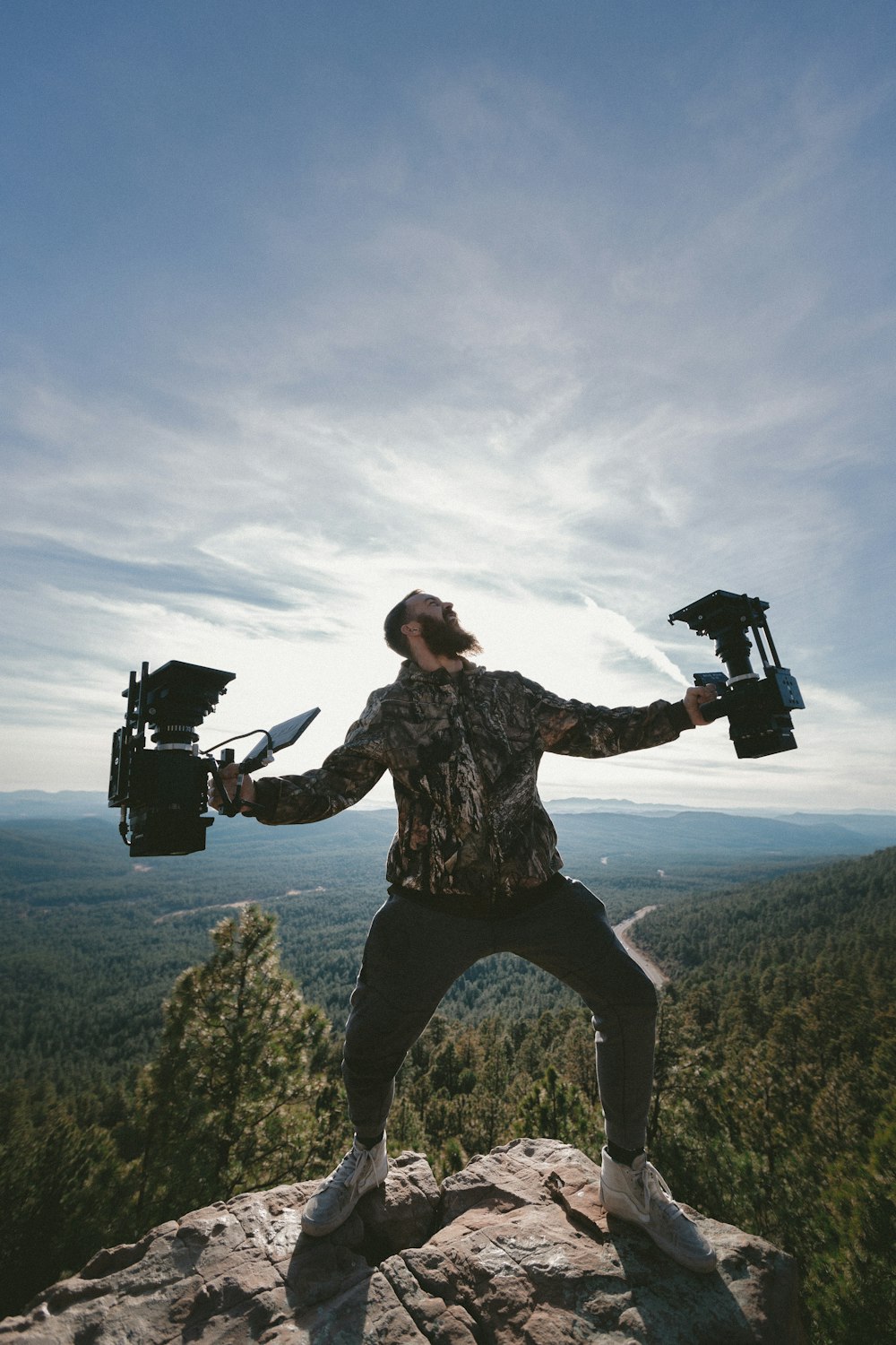 person standing on rocky mountain peak while holding two cameras