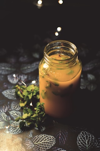A glass jar filled with a creamy orange beverage, garnished with fresh herbs, sits on a dark, textured surface adorned with leaf patterns. A sprig of green mint lies beside the jar, with soft, warm fairy lights illuminating the scene, creating a cozy ambiance.