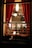 A cozy dining scene viewed through a window, showcasing people gathered around tables in a warmly lit restaurant. The interior features red curtains and pendant lighting that create an intimate atmosphere.