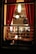 A cozy dining scene viewed through a window, showcasing people gathered around tables in a warmly lit restaurant. The interior features red curtains and pendant lighting that create an intimate atmosphere.