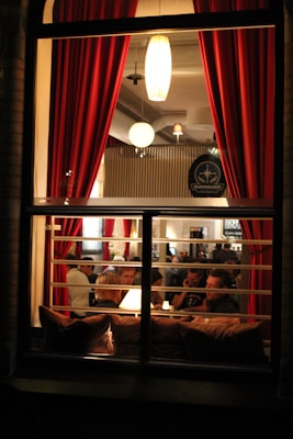 A cozy dining scene viewed through a window, showcasing people gathered around tables in a warmly lit restaurant. The interior features red curtains and pendant lighting that create an intimate atmosphere.