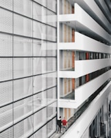 A modern interior space with a large glass facade on the left and an open corridor on the right. The corridor is lined with doors and several exit signs. Two people are walking along the corridor, providing a sense of scale. The design features clean lines and a minimalist aesthetic.