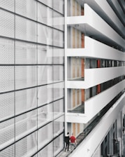 A modern interior space with a large glass facade on the left and an open corridor on the right. The corridor is lined with doors and several exit signs. Two people are walking along the corridor, providing a sense of scale. The design features clean lines and a minimalist aesthetic.