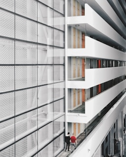 A modern interior space with a large glass facade on the left and an open corridor on the right. The corridor is lined with doors and several exit signs. Two people are walking along the corridor, providing a sense of scale. The design features clean lines and a minimalist aesthetic.