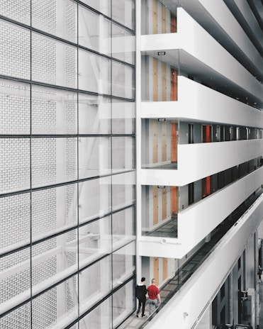 A modern interior space with a large glass facade on the left and an open corridor on the right. The corridor is lined with doors and several exit signs. Two people are walking along the corridor, providing a sense of scale. The design features clean lines and a minimalist aesthetic.