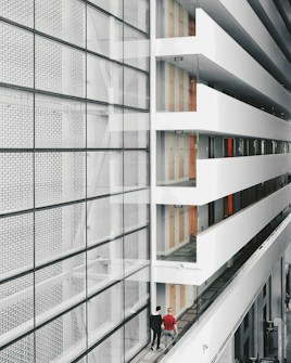 A modern interior space with a large glass facade on the left and an open corridor on the right. The corridor is lined with doors and several exit signs. Two people are walking along the corridor, providing a sense of scale. The design features clean lines and a minimalist aesthetic.