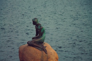 A bronze statue of a mermaid sits on a large rock near a body of water. The water is slightly choppy, with a bluish-green tint. The mermaid is depicted in a seated position, appearing contemplative.