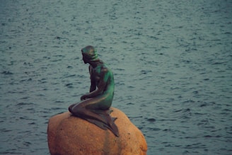 A bronze statue of a mermaid sits on a large rock near a body of water. The water is slightly choppy, with a bluish-green tint. The mermaid is depicted in a seated position, appearing contemplative.
