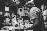 People are gathered around a table, eating and interacting energetically. It appears to be in a casual, possibly industrial or makeshift setting, with visible lighting fixtures and a partially structured ceiling. The atmosphere is lively and informal.