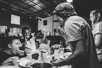 People are gathered around a table, eating and interacting energetically. It appears to be in a casual, possibly industrial or makeshift setting, with visible lighting fixtures and a partially structured ceiling. The atmosphere is lively and informal.