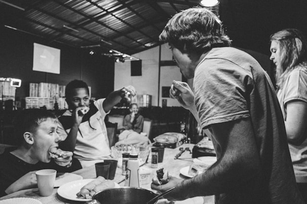 People are gathered around a table, eating and interacting energetically. It appears to be in a casual, possibly industrial or makeshift setting, with visible lighting fixtures and a partially structured ceiling. The atmosphere is lively and informal.