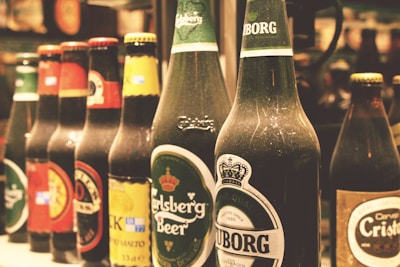 A row of various beer bottles with distinct labels, including brands such as Carlsberg and Tuborg. The bottles are arranged side by side on a shelf with a slightly blurred background, indicating a bar or store setting.