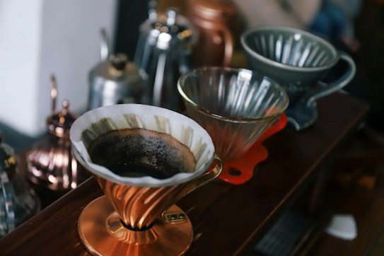 A wooden surface holds several pour-over coffee drippers, one of which is in use with a paper filter and freshly brewed coffee grounds. The drippers are made of various materials, including metal and ceramic, and some have intricate designs. A collection of metal kettles is blurred in the background.