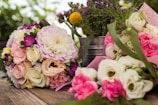 A variety of colorful floral arrangements featuring roses, peonies, and other blossoms are placed on a wooden surface. The bouquets have hues of pink, white, yellow, and purple, complemented by green leaves and stems. A metallic bucket in the background holds additional flowers, adding depth to the scene.