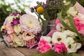 A variety of colorful floral arrangements featuring roses, peonies, and other blossoms are placed on a wooden surface. The bouquets have hues of pink, white, yellow, and purple, complemented by green leaves and stems. A metallic bucket in the background holds additional flowers, adding depth to the scene.