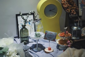 A decorative setup featuring a large yellow clock, positioned prominently against the backdrop. In the foreground are various decorative items including a green glass vase with eucalyptus, a small clear glass dome with a ribbon, and a card with floral designs resting on a wooden base. To the right, a tiered stand holds dried flowers, and off to the side, a metallic lantern adds a rustic touch. The setup is arranged on a soft lavender cloth, enhancing the calming atmosphere.