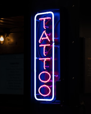 A vertical neon sign displaying the word 'TATTOO' in vibrant pink and blue hues, mounted against a dark background. The letters are outlined with a blue neon border that contrasts sharply, giving a modern and urban feel to the environment.