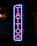 A vertical neon sign displaying the word 'TATTOO' in vibrant pink and blue hues, mounted against a dark background. The letters are outlined with a blue neon border that contrasts sharply, giving a modern and urban feel to the environment.