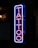 A vertical neon sign displaying the word 'TATTOO' in vibrant pink and blue hues, mounted against a dark background. The letters are outlined with a blue neon border that contrasts sharply, giving a modern and urban feel to the environment.