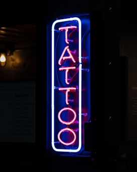 A vertical neon sign displaying the word 'TATTOO' in vibrant pink and blue hues, mounted against a dark background. The letters are outlined with a blue neon border that contrasts sharply, giving a modern and urban feel to the environment.