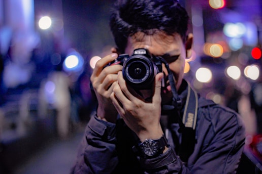 A person holding a Nikon DSLR camera close to their face, focusing on taking a photo. The background is blurred with colorful lights creating a bokeh effect, likely indicating an urban nighttime setting. The person is wearing dark clothing and has a black watch on their wrist.