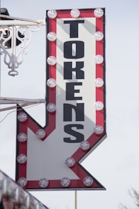 A vintage-style illuminated sign with the word 'TOKENS' in bold black letters. The letters are arranged vertically on a white background, bordered with red, with round lights outlining the edges of the sign. The bottom of the sign forms an arrow pointing downward.
