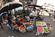 A vibrant outdoor market scene with several people interacting and trading goods. Various colorful fruits and flowers are displayed in baskets and crates on the ground and tables. An assortment of umbrellas provides shade to the vendors and customers. The surrounding architecture appears traditional and there is a bustling atmosphere.