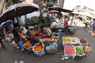 A vibrant outdoor market scene with several people interacting and trading goods. Various colorful fruits and flowers are displayed in baskets and crates on the ground and tables. An assortment of umbrellas provides shade to the vendors and customers. The surrounding architecture appears traditional and there is a bustling atmosphere.