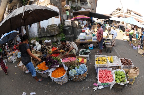 A vibrant outdoor market scene with several people interacting and trading goods. Various colorful fruits and flowers are displayed in baskets and crates on the ground and tables. An assortment of umbrellas provides shade to the vendors and customers. The surrounding architecture appears traditional and there is a bustling atmosphere.