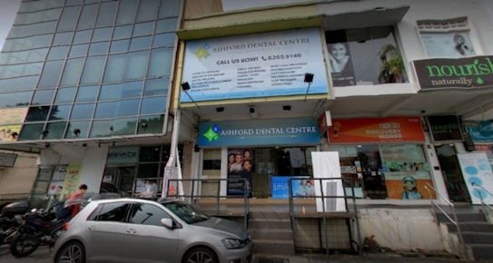 The image depicts a street view of a building with various businesses. A large sign for Ashford Dental Centre is prominently displayed above the entrance. Several other signs are visible, including one for a place named 'nourish naturally.' There is a parked car and a few motorcycles in front of the building, and a couple of pedestrians are nearby.