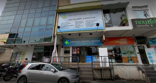 The image depicts a street view of a building with various businesses. A large sign for Ashford Dental Centre is prominently displayed above the entrance. Several other signs are visible, including one for a place named 'nourish naturally.' There is a parked car and a few motorcycles in front of the building, and a couple of pedestrians are nearby.