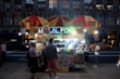 A bustling street food vendor at night with a brightly lit sign that reads 'HALAL FOOD'. The stall is adorned with colorful images of various food items and is covered by two yellow and red umbrellas. Several people stand around, appearing to buy or receive food, while a taxi and other vehicles are visible in the background on a busy street.