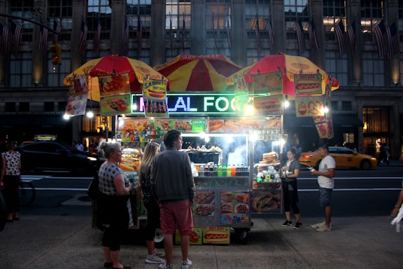 A bustling street food vendor at night with a brightly lit sign that reads 'HALAL FOOD'. The stall is adorned with colorful images of various food items and is covered by two yellow and red umbrellas. Several people stand around, appearing to buy or receive food, while a taxi and other vehicles are visible in the background on a busy street.