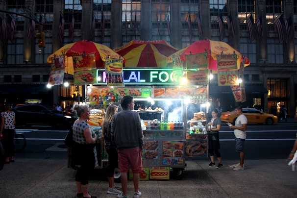 A bustling street food vendor at night with a brightly lit sign that reads 'HALAL FOOD'. The stall is adorned with colorful images of various food items and is covered by two yellow and red umbrellas. Several people stand around, appearing to buy or receive food, while a taxi and other vehicles are visible in the background on a busy street.