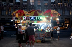 A bustling street food vendor at night with a brightly lit sign that reads 'HALAL FOOD'. The stall is adorned with colorful images of various food items and is covered by two yellow and red umbrellas. Several people stand around, appearing to buy or receive food, while a taxi and other vehicles are visible in the background on a busy street.